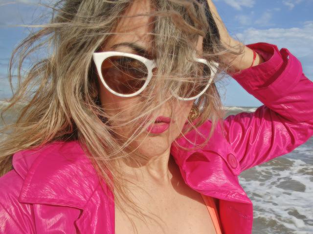 Maytena with messy blonde hair, white cat-eye sunglasses, pink glossy lips, and a shiny pink jacket, holding her hair up, standing by the ocean, showing her tits.