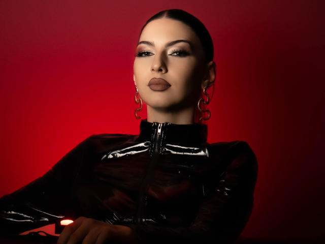 LucyGrace with slicked-back hair, wearing a shiny black latex top, hoop earrings, and dark lipstick, looking seductively at the camera against a red background.