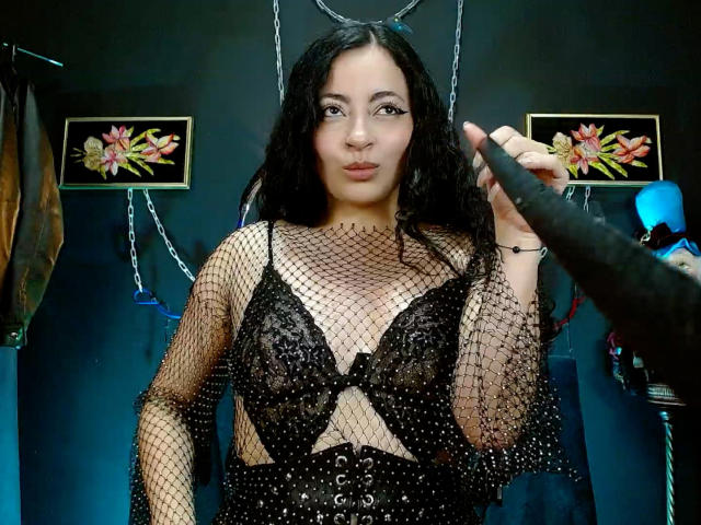 LunaTikka with black curly hair, wearing a see-through fishnet top over black lace bra, holding a black dildo in front of her, pouting her lips.