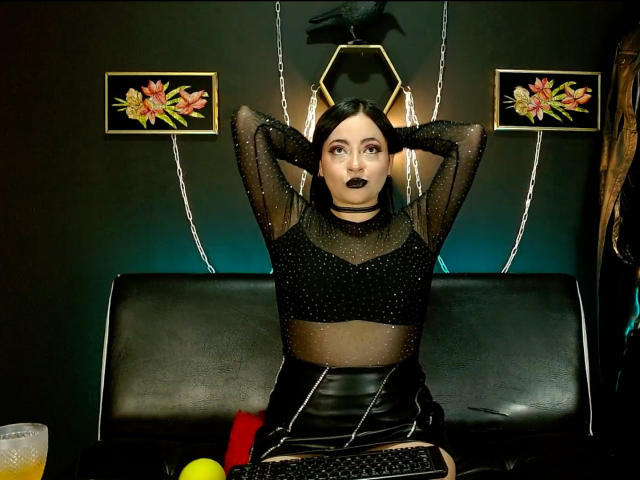 LunaTikka with dark lipstick, black sheer top, and black leather skirt, hands behind head, sitting on black bench, surrounded by flowers, orange drink, and yellow ball.