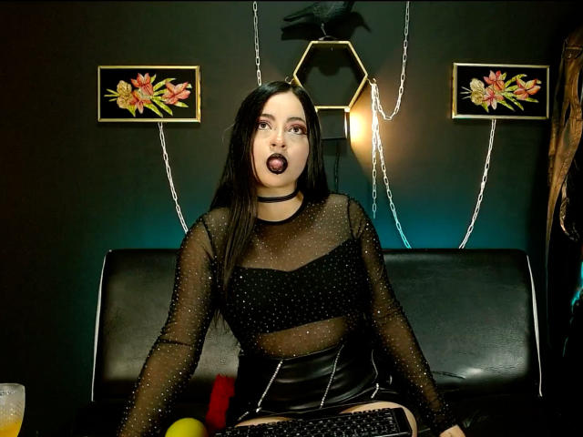 LunaTikka, wearing a sheer black top with sparkles and a black choker, has dark lipstick and long black hair, sitting on a black leather couch with a black skirt.