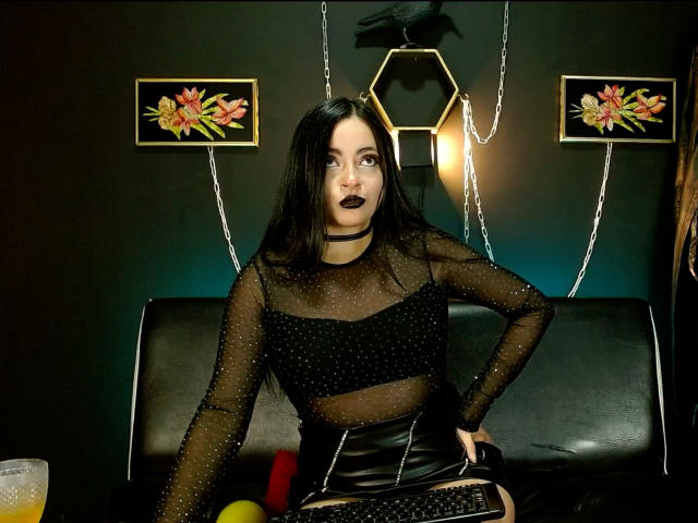 LunaTikka with black goth makeup, sheer polka-dot top, black leather skirt, sitting on black couch, dark hair, black choker, black lipstick, neon hexagon light, floral paintings, glass with orange drink.