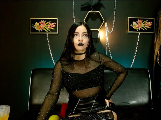 LunaTikka with black hair, goth makeup, black sheer top, black polka-dot bra, black leather skirt, sitting on black couch, hands on hips, dark lipstick, goth decor, orange drink.
