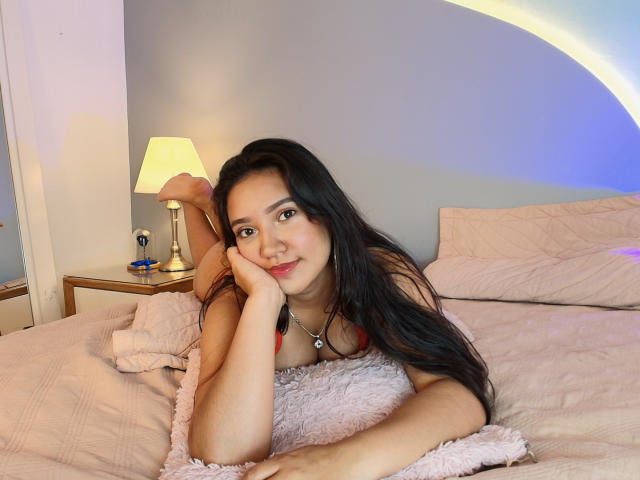 LaylaMoon with long black hair, laying on a bed, smiling, head on hand, wearing a red bra, pink pillow, lamp on nightstand, gray wall, soft blue light.