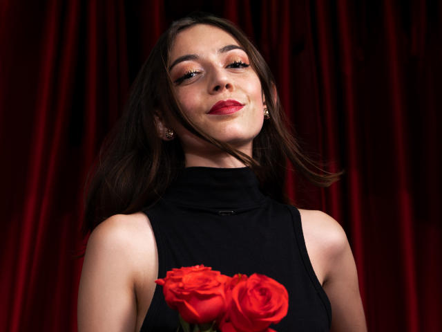 VictoriaPalmerfg stands against a dark red velvet backdrop. She has fair skin, dark brown hair, and wears red lipstick. She's dressed in a black sleeveless top with a high neck. She holds three red roses in front of her. Her eyes are slightly closed, and she has a subtle, confident smile. Her earrings are small and round. The red velvet background contrasts with her black top and the bright red roses. The composition focuses on her upper body and face.