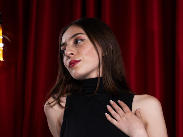 VictoriaPalmerfg with long brown hair, red lipstick, black sleeveless turtleneck, hand on chest, looking to the side, red curtain background, soft light, pale skin, subtle makeup.