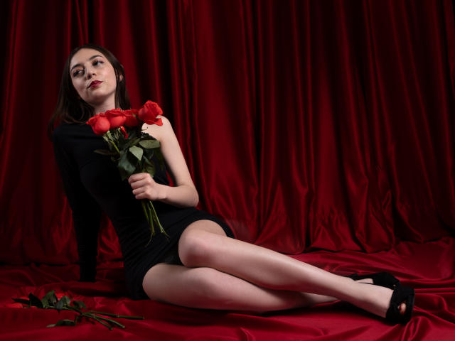 VictoriaPalmerfg, with pale skin and dark hair, wearing a black dress, holding red roses, sitting on red satin, legs crossed, black heels, looking at camera, rose on floor.