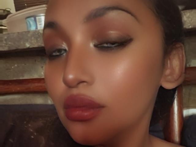 Close-up of MckenzieOne's face with brown skin and glossy makeup. She has dark, arched eyebrows, subtle eyeshadow, and black eyeliner that extends into a cat-eye style. Her full, red-painted lips are slightly parted. Her eyes are half-closed, giving a relaxed expression. The background is blurry, featuring a concrete surface and a wooden shelf. The lighting is soft, highlighting her smooth skin and subtle shine.