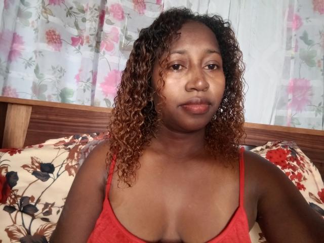 LuciaSexy69, dark-skinned woman with curly brown hair, red tank top, small tits, sitting on bed with floral pillow, looking at camera with neutral expression.