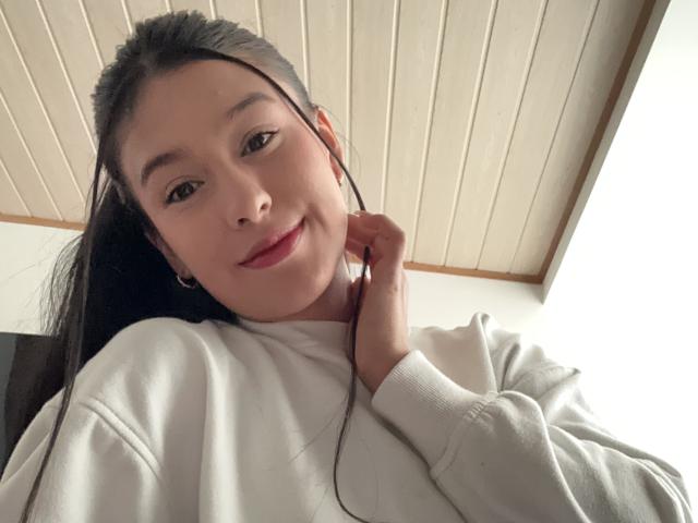 Soranaa, con lunghi capelli neri, pelle chiara e rossetto rosso, indossa un maglione bianco, sorride leggermente, ha le cuffie nelle orecchie, una mano vicino alla guancia, sullo sfondo si vede un soffitto con pannelli di legno..