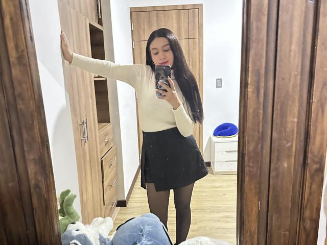 Soranaa met lang zwart haar, een witte top met lange mouwen, een zwarte rok, zwarte leggings, staat voor een spiegel en maakt een selfie. Links is een houten kast te zien, rechts een blauw kussen..