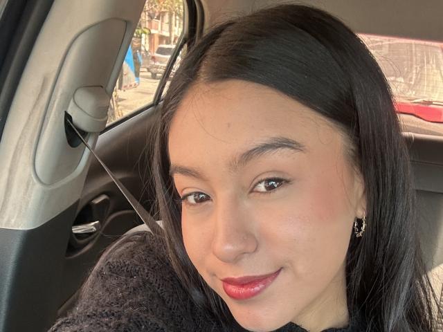 Soranaa's face in a car, with black hair, brown eyes, red lipstick, black sweater, and gold earrings, smiling slightly. Her ass is not visible.