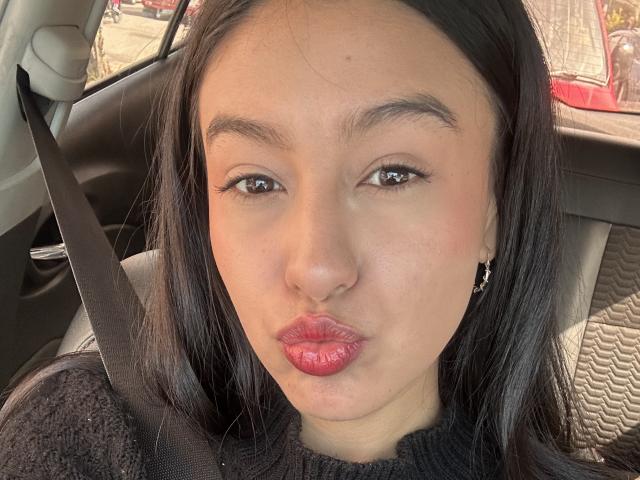 Los labios fruncidos y pintados de rojo de Soranaa están haciendo una mueca dentro de un coche, con un cinturón de seguridad sobre su cabeza de pelo negro y un suéter negro puesto..
