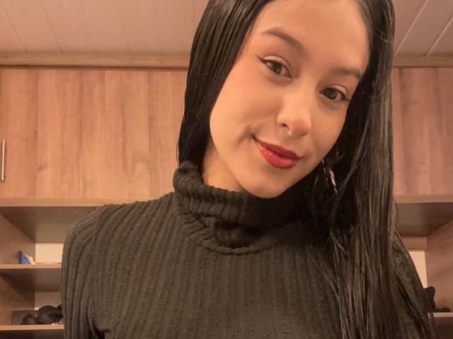 Soranaa, con cabello negro largo, vestida con un cuello alto negro texturizado, lápiz labial rojo, sonriendo y con un pequeño piercing en la nariz, en una habitación de madera..