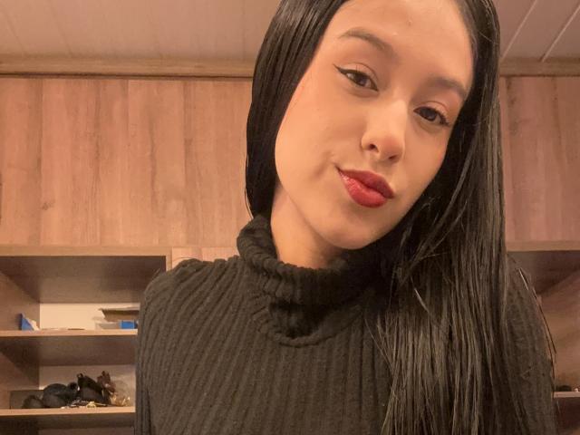 Soranaa, con cabello negro largo, vestida con un cuello alto negro texturizado, lápiz labial rojo y delineador de ojos alado, sonriendo ligeramente, mostrando su nariz y parte superior del pecho..