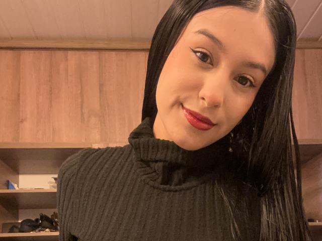 Soranaa, con i suoi lunghi capelli neri, il rossetto rosso e il maglione nero a coste, sorride leggermente alla fotocamera. I suoi occhi marroni sono contornati da eyeliner nero..