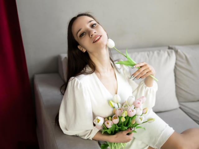StacieLove with long brown hair, white blouse, holding bouquet of pink and white tulips, sitting on gray sofa, holding one tulip to mouth, smiling.