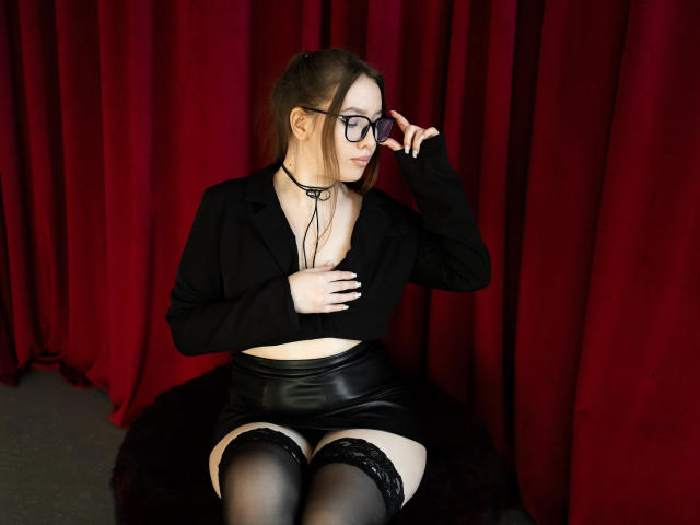 DanielleSweetty sits on a black furry surface against red curtains. She wears a black long-sleeve crop top with a deep V-neck, black leather skirt, and thigh-high black stockings with lace tops. She has fair skin, brown hair in a half-up style, and wears black-framed glasses. She touches her glasses with her right hand, looking to the left. Her left hand is placed on her chest. The image uses a realistic, photographic style.