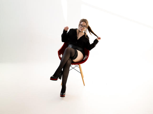 DanielleSweetty with glasses, black blouse, short skirt, thigh-high stockings, black heels, red chair, holding her ponytail, white background, slightly smiling, legs crossed.