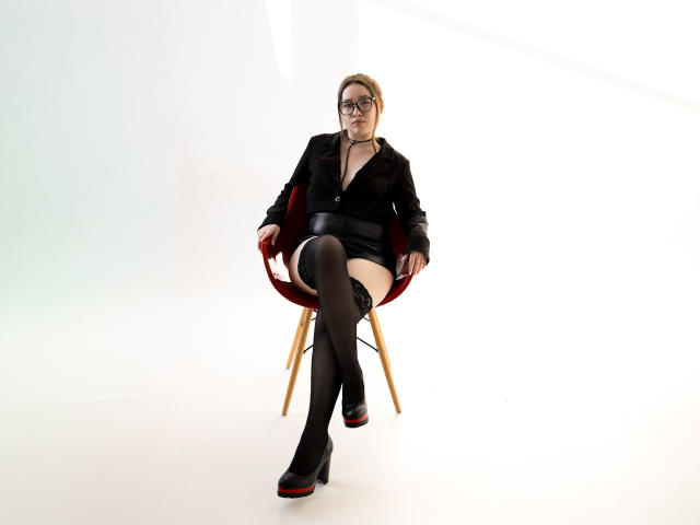 DanielleSweetty sits in a red mid-century modern chair with wooden legs. She wears a black button-up blouse, black mini skirt, black thigh-high stockings, and black platform heels with red soles. She has blonde hair, glasses, and a confident expression. Her legs are crossed, showing a white thigh. The photograph uses bright, even lighting, highlighting her dark clothing and the chair's red seat. The image focuses on her stylish, slightly provocative outfit.