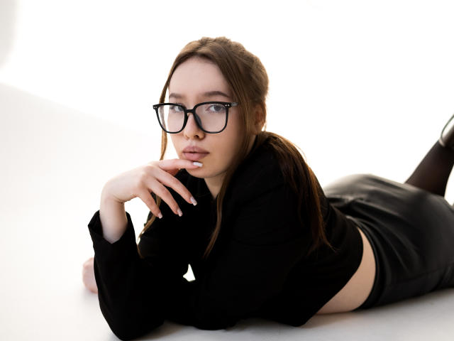 DanielleSweetty lies on her stomach with her head propped up by her right hand. She has light skin, brown hair in a ponytail, and wears black-rimmed glasses. She has a serious expression with slightly parted lips. She's dressed in a black long-sleeve top and black leather pants. Her left arm is bent, and her right hand rests on her chin. The white background is minimal, focusing on her. The composition is sharp with a clean, bright look. Her nails are long and manicured. The image has a modern, stylish vibe.