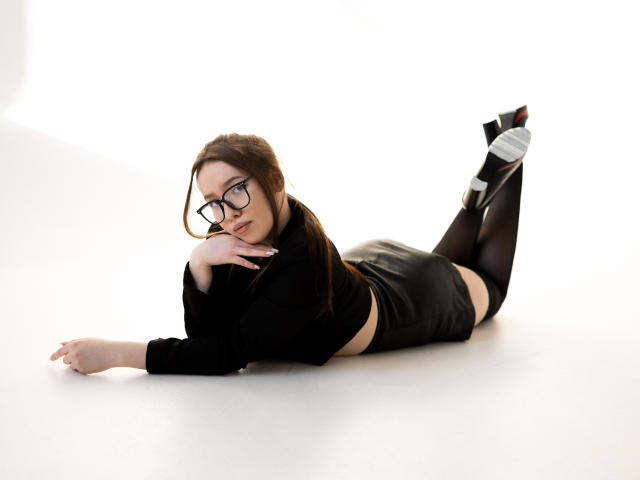 DanielleSweetty, lying on her stomach, black crop top, black leather skirt, black thigh-highs, glasses, looking at camera, hands under chin, high heels, white background, butt up.