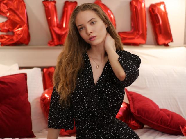AlicePearl with long blonde hair, wearing a black polka dot dress, has a cute face and small tits, sitting on a bed with red pillows and shiny red letters.