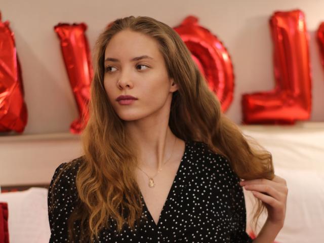 AlicePearl with long, wavy brown hair, wearing a black polka dot shirt, looking to the side with a slight pout, holding her hair, gold necklace, red balloon background.