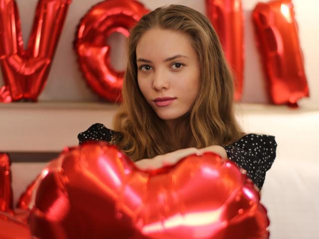 AlicePearl with long, wavy brown hair, wearing a black polka-dot top, holding shiny red heart balloons, looking at the camera with a subtle smile, soft light.