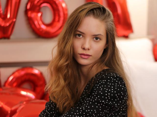 AlicePearl with long, wavy, blonde hair, wearing a black, polka-dot top, has a serious, slightly pouty expression, and red balloon letters are in the background.