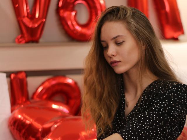 AlicePearl with long, wavy brown hair, wearing a black polka dot top, looking down with a serious expression, red balloon letters in the background.