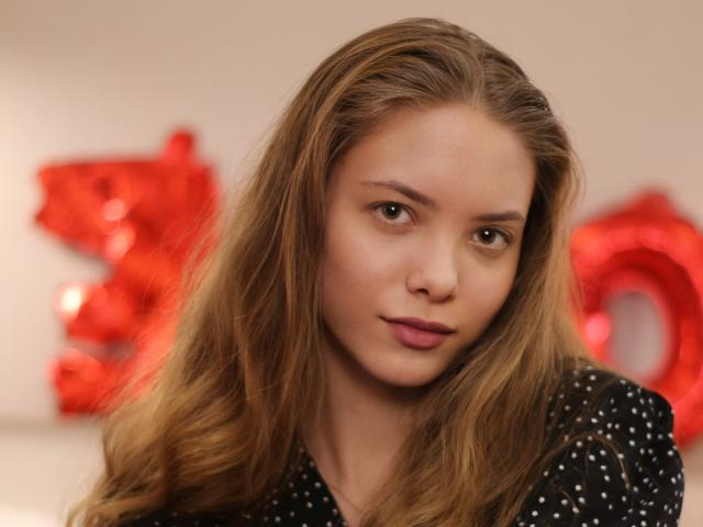 AlicePearl has long, wavy brown hair, light skin, and brown eyes. She's wearing a black polka-dot shirt, with a slight pout and soft, natural makeup.