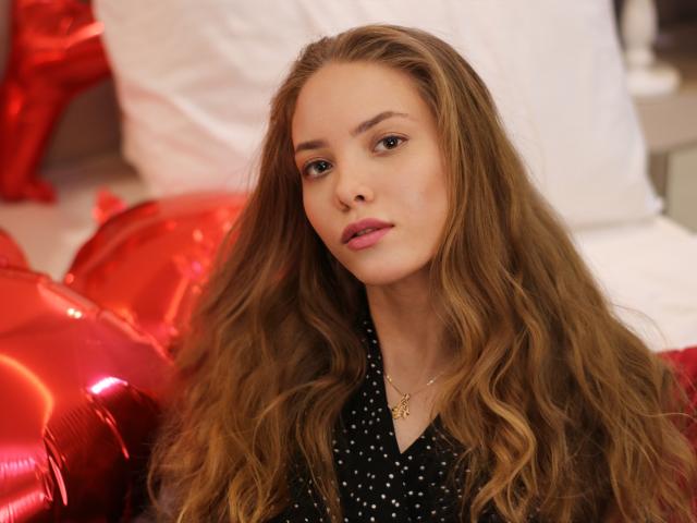 AlicePearl with long wavy brown hair, pink lipstick, black polka dot shirt, gold necklace, and red shiny balloons in the background, looking slightly bored.