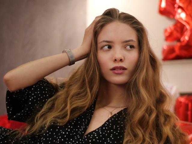 AlicePearl has long, wavy brown hair, wearing a black polka dot dress, looking to the side with her hand in her hair, a silver bracelet, and pink lipstick.