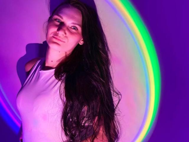 ErrosAy with long dark hair, white shirt, small tits, and a slight smile, standing against colorful light swirls, purple and green hues, soft lighting, gentle expression.