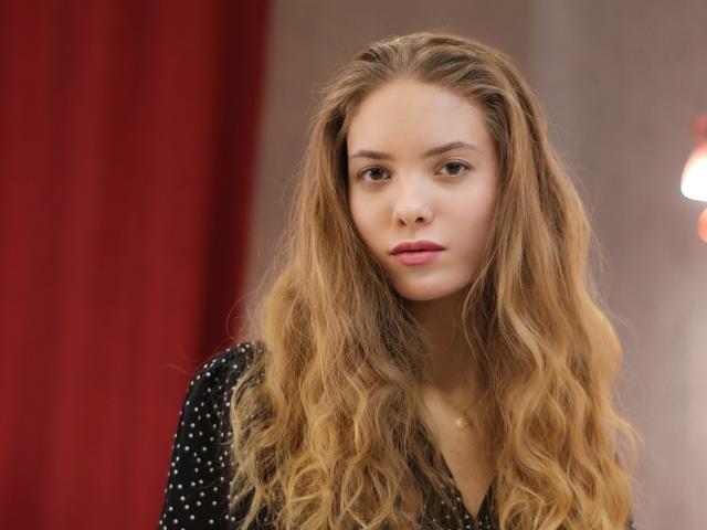 AlicePearl with long wavy blonde hair, wearing a black polka dot top, has a serious expression, pink lips, and a small gold necklace, in a well-lit indoor setting.