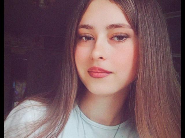 Close-up portrait of KeylasRebecca. She has long, straight brown hair, slightly parted in the middle, covering her shoulders. Her skin is fair with a smooth texture. She has light brown eyes with subtle eyeliner and full pink lips. KeylasRebecca wears a light grey shirt. The lighting is soft, creating gentle shadows on her face. The image has a slightly warm, digital filter. Her expression is neutral, slightly smiling. The composition focuses on her face, with her hair framing her face on both sides.