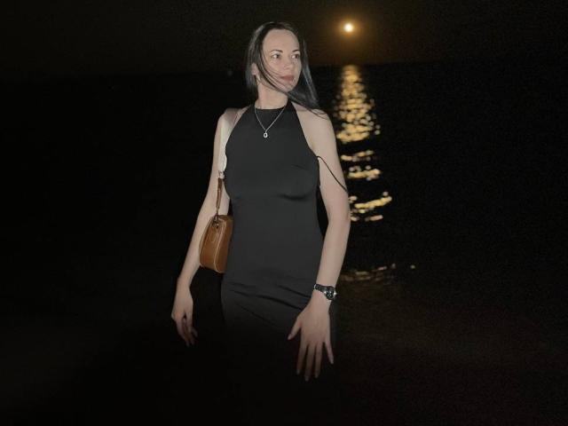 Dalaila with long black hair, black sleeveless dress, brown leather bag, necklace, and wristwatch, standing by dark water with moonlight reflection. Her tits are small, she's smiling.