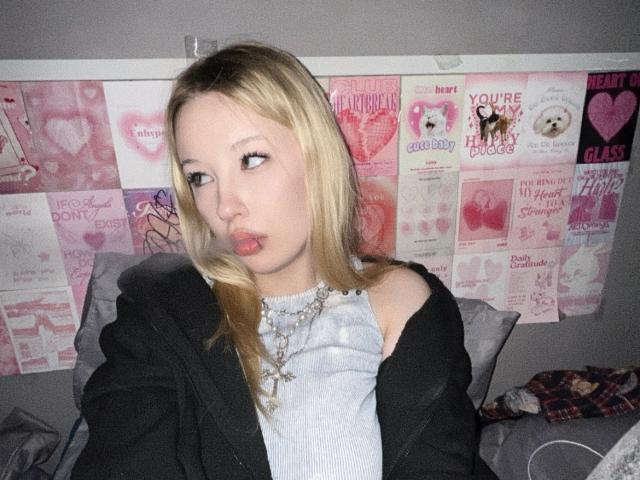 StarSice with blonde hair, pouty lips, wearing a black jacket over a grey top, necklace, looking to the side, in a bedroom with pink posters on the wall.