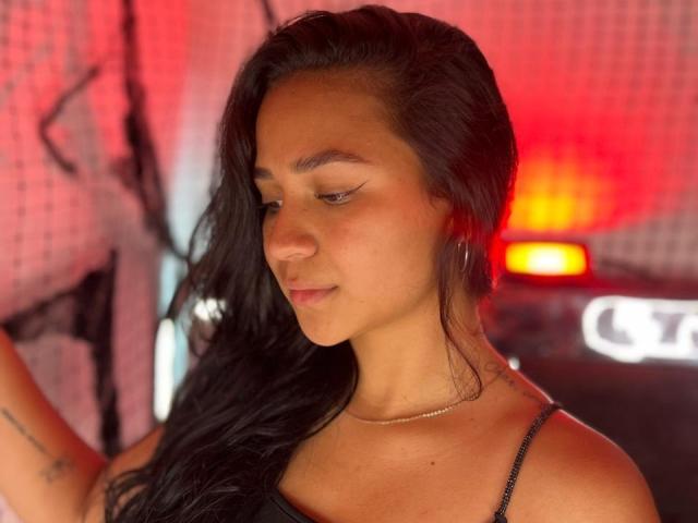 RouxiMori with long black hair, brown skin, subtle makeup, and a thin black tank top, wearing a gold chain necklace, standing with a red light in the background.