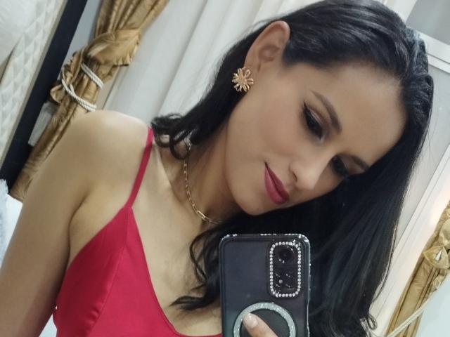 VanesaValet with black hair, red dress, gold star earrings, and pink lipstick, holding phone, showing her tits in mirror selfie. She's looking down, smiling slightly.