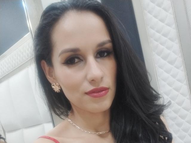 VanesaValet with long black hair, red lipstick, and a gold chain necklace, wearing floral earrings, looking at the camera with a slight smile. Boobs visible under red top.