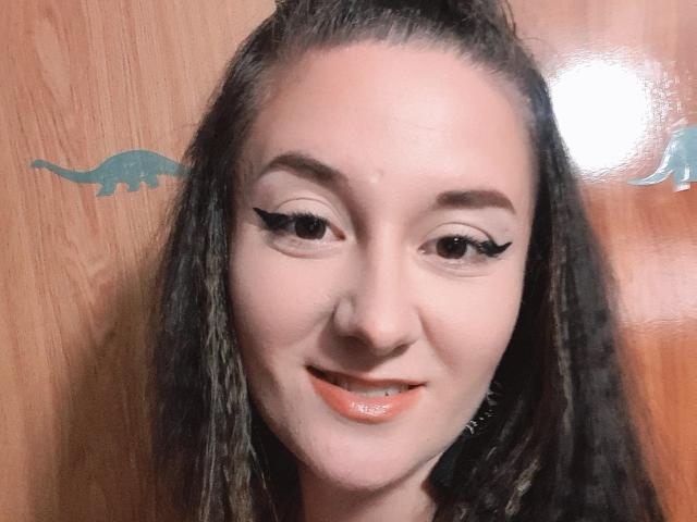 Close-up of LexyAlexa with fair skin, dark brown hair in a top knot, and long, wavy strands. She has black eyeliner with a sharp, winged tip on her upper eyelids. Her brown eyes are slightly squinted, and she's smiling with glossy, orange-pink lipstick. The wooden background has a small, green dinosaur sticker on the left. Light reflects off the right side of her face. Her hair has a slight texture with visible strands.