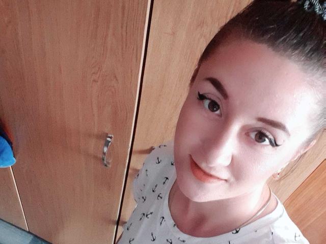 LexyAlexa with light skin, brown hair in a bun, wearing a white shirt with small black prints, has black eyeliner and a slight smile, standing in front of wooden cabinets.