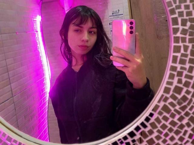 Katherin Johns, con i capelli scuri e ondulati, indossa una giacca nera con zip e sta scattando un selfie allo specchio con una luce al neon rosa, tenendo un iPhone rosa, con il suo riflesso visibile in uno specchio a rete bianco..