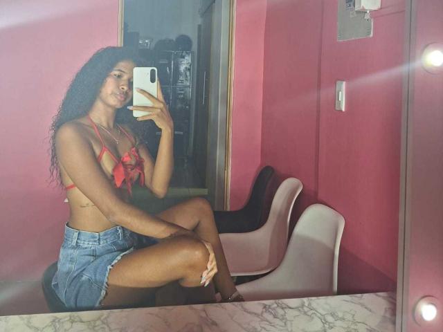 Tifannyy with long curly black hair, red bra with a bow, showing her tits, and blue denim shorts, sitting on a chair, taking a mirror selfie.