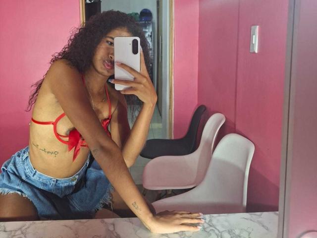 Tifannyy with curly dark hair, wearing a red bra showing small tits and denim shorts, taking a mirror selfie with a white phone, pouty lips, tattoo on ribcage.