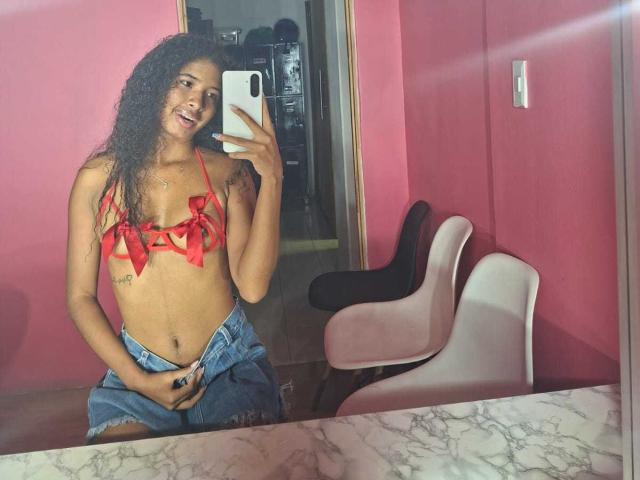 Tifannyy with curly hair, red ribbon bikini top, small tits, and blue denim shorts, smiling at mirror selfie, holding phone, pink room, white chair, black chair.