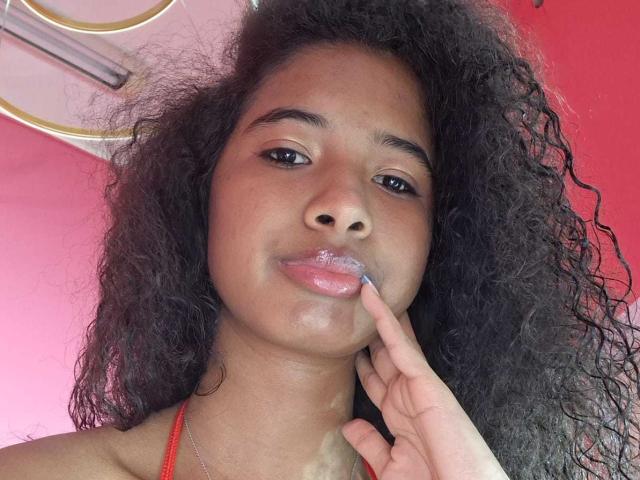 Tifannyy with curly black hair, brown skin, and glossy pink lips, applying blue lipstick with her right finger, wearing a red halter top, smiling softly.