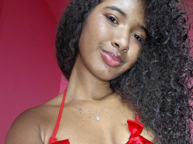 Tifannyy with curly black hair, brown skin, and pink lips, wearing a red lace bra with bows, and a silver necklace, smiling at the camera. Boobs visible.